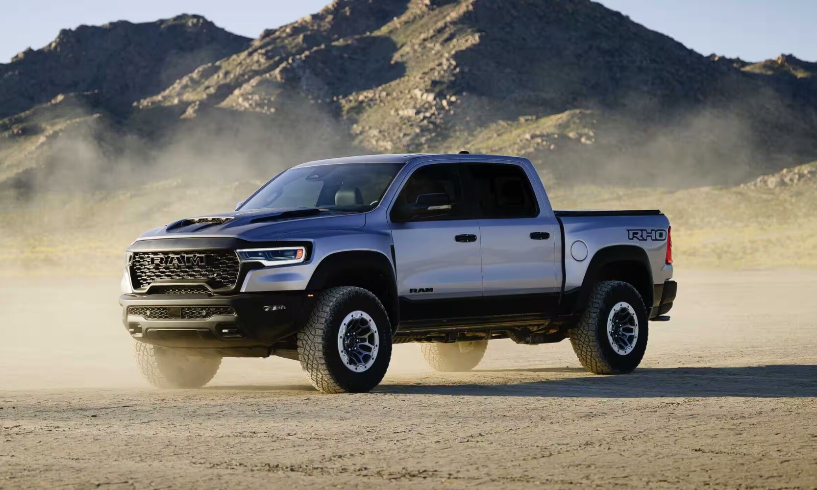 2025%20GMC%20Sierra%201500%20vs.%202025%20RAM%201500%20-%202025%20RAM%201500%20RHO%20-%20Octane%20GMC%20of%20Santa%20Fe.jpg Exterior side image of a white 2025 RAM 1500 RHO parked in a dirt field with mountains in the background