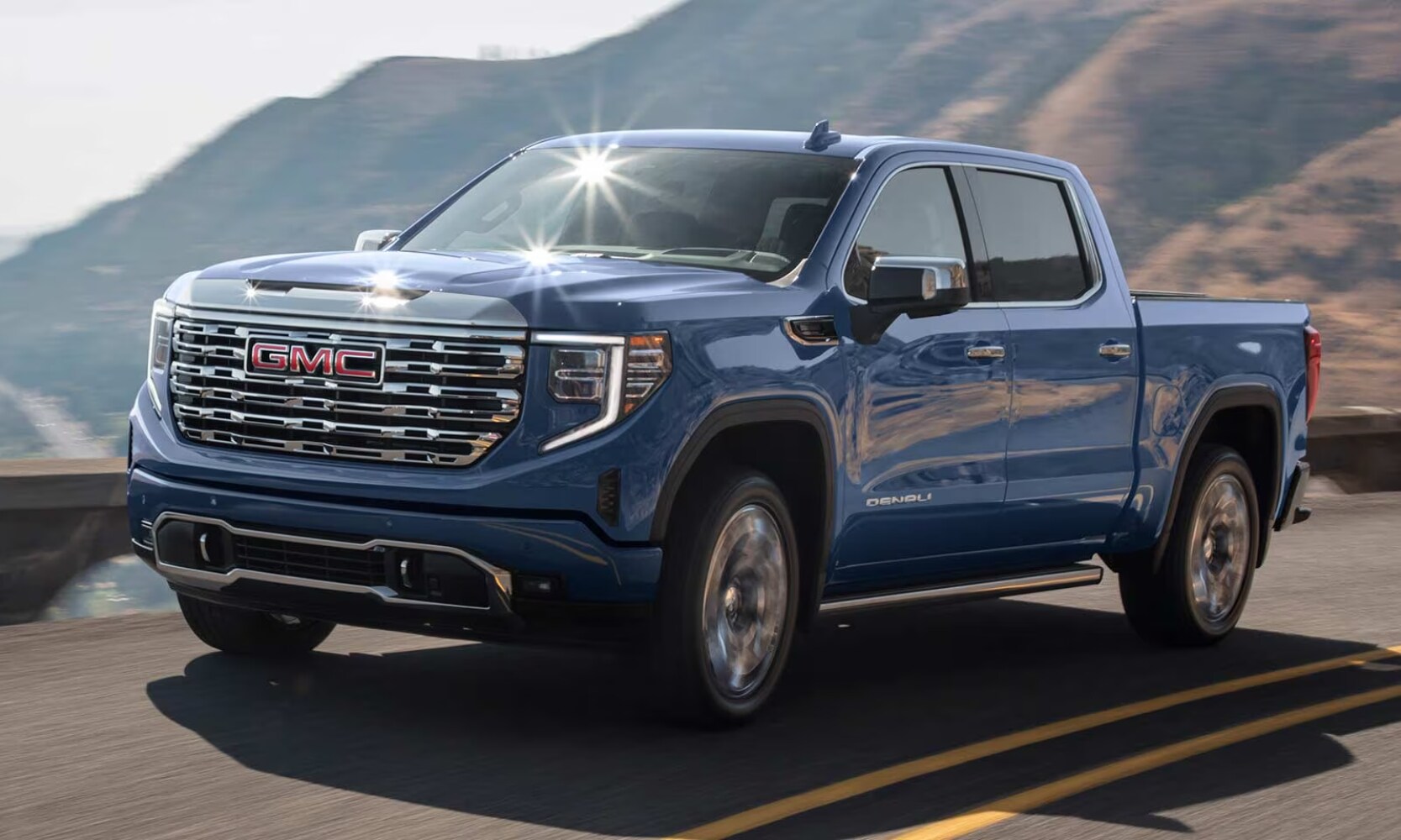 2025%20GMC%20Sierra%201500%20vs.%202025%20RAM%201500%20-%202025%20GMC%20Sierra%201500%20Denali%20-%20Octane%20GMC%20of%20Santa%20Fe.jpg Exterior side angle image of a blue 2025 GMC Sierra 1500 Denali driving on a highway with mountains in the background