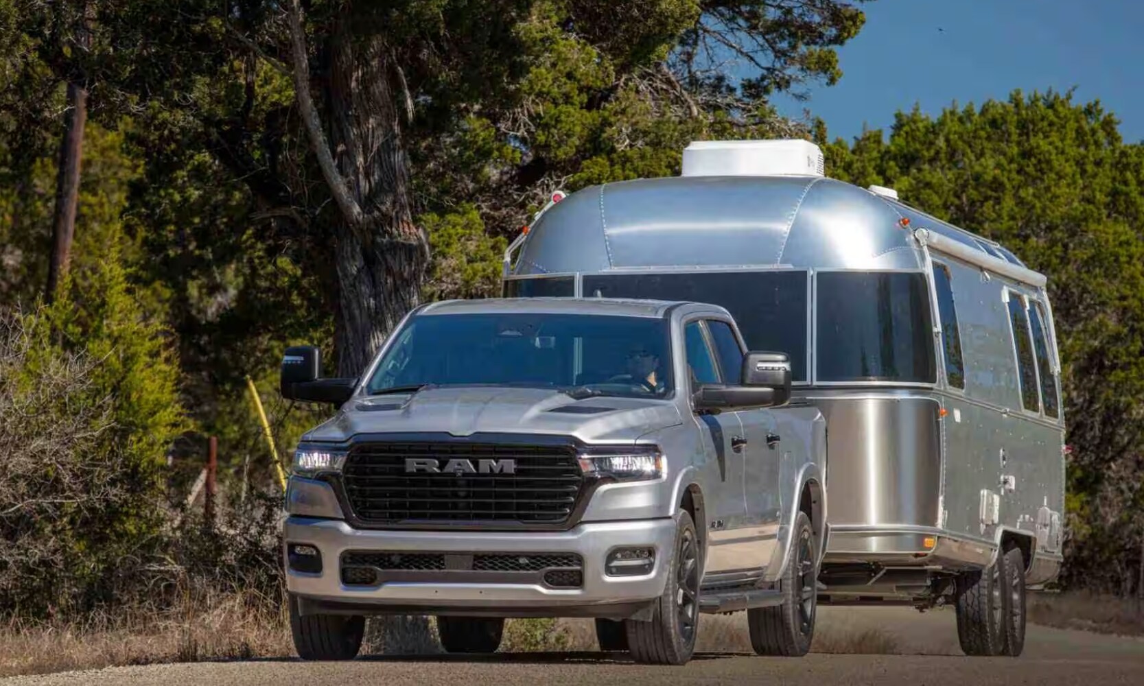2025%20GMC%20Sierra%201500%20vs.%202025%20RAM%201500%20-%202025%20RAM%201500%20Towing%20Comparison%20-%20Octane%20GMC%20of%20Santa%20Fe.jpg Exterior front image of a silver 2025 RAM 1500 towing a camping trailer on a road with trees in the background