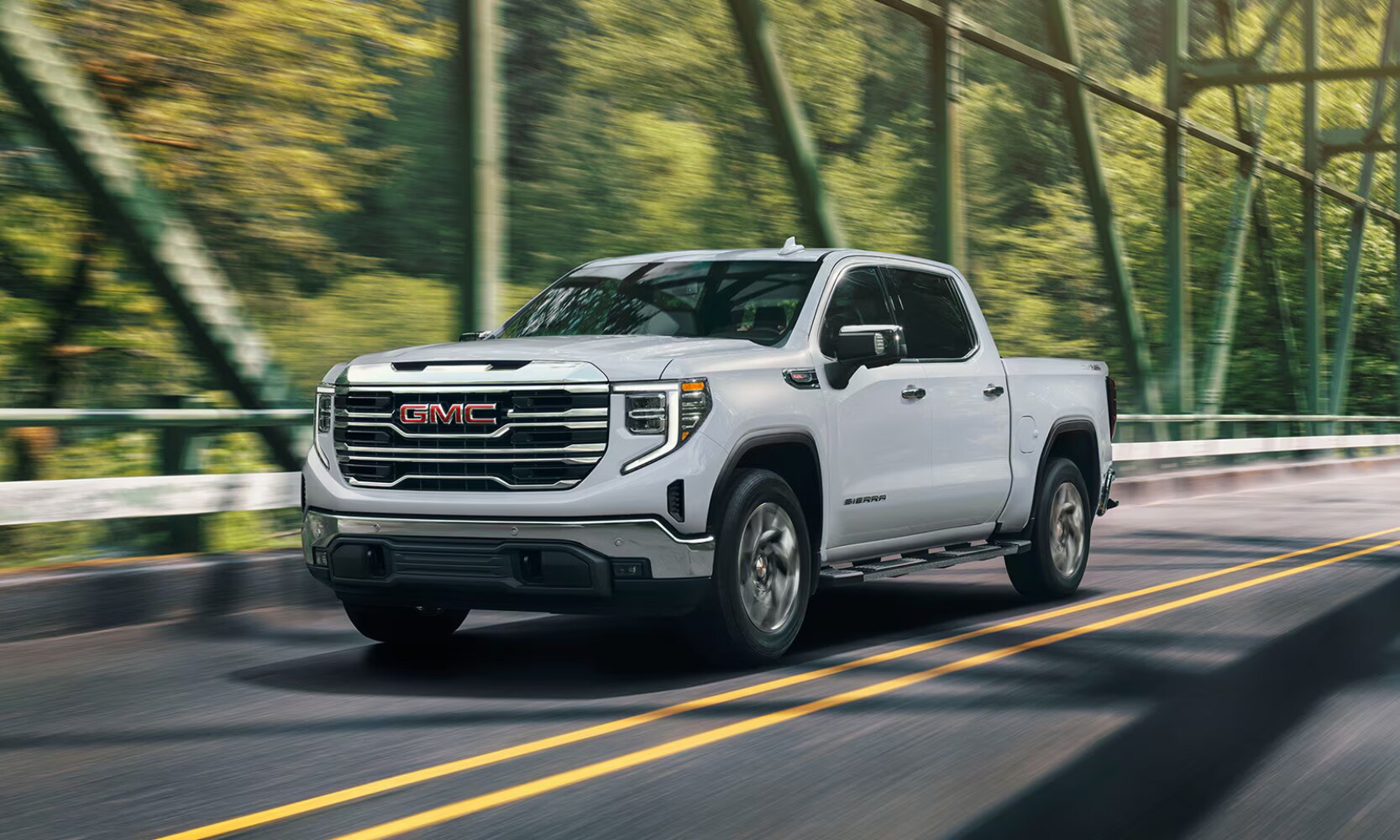 Exterior front angle hero image of a white 2026 GMC Sierra 1500 driving over a tree-lined bridge