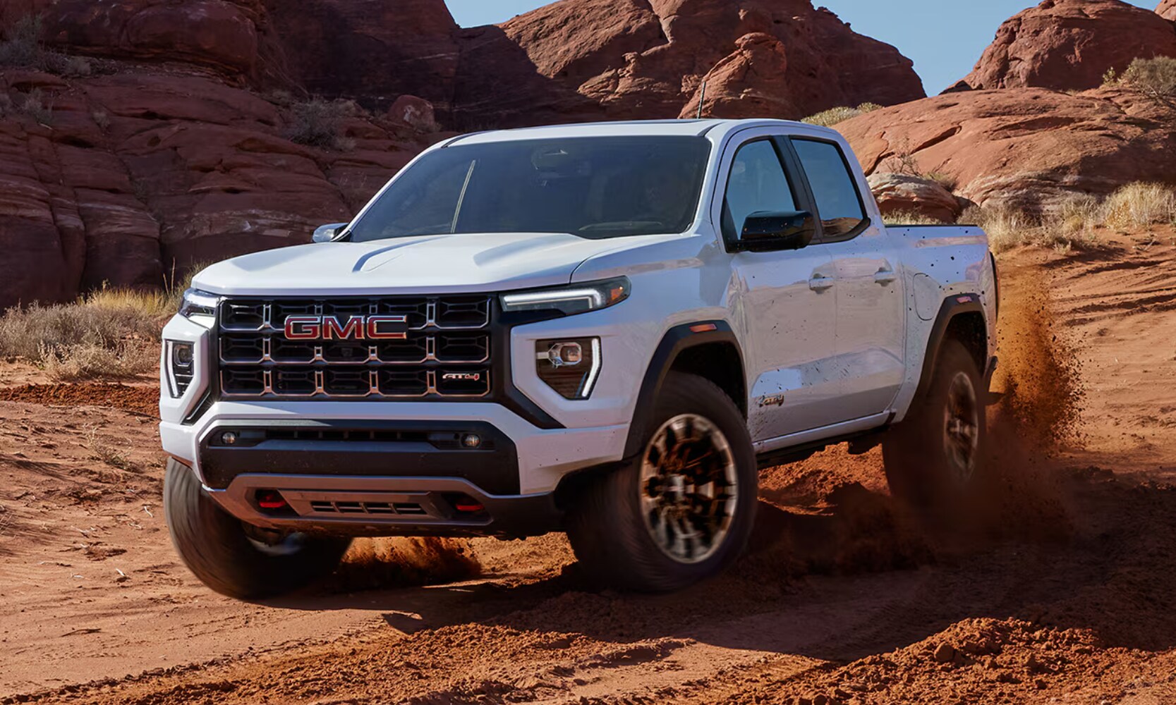 Exterior front angle image of a white 2026 GMC Canyon AT4 off-roading through dirt in a rocky mountainous landscape