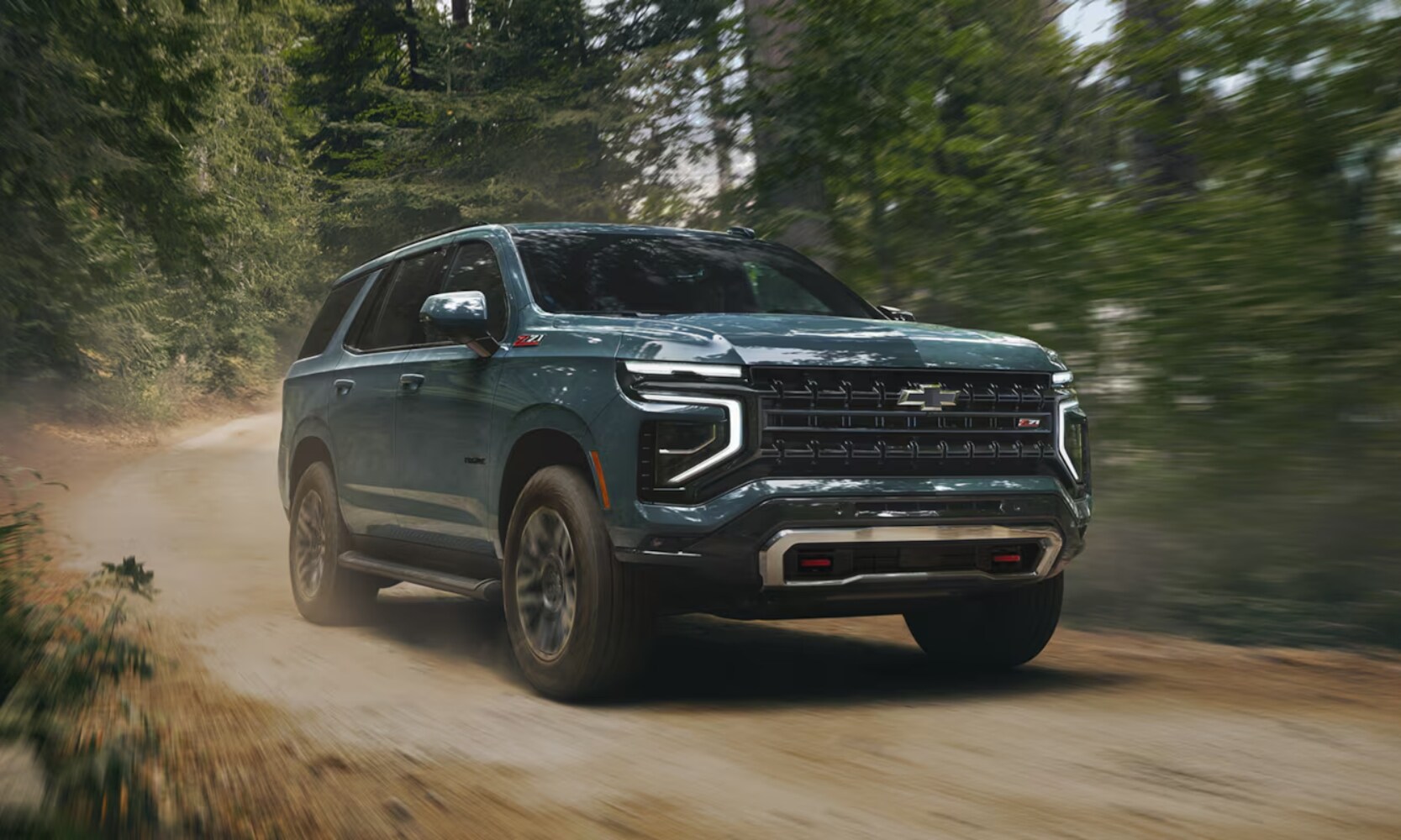 2025%20GMC%20Yukon%20vs.%202025%20Chevrolet%20Tahoe%20-%202025%20Chevy%20Tahoe%20Z71%20-%20Octane%20GMC%20of%20Santa%20Fe.jpg Exterior front angle image of a teal 2025 Chevy Tahoe Z71 driving on a dirt road with surrounding trees