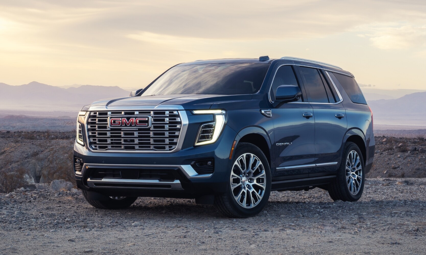 2025 GMC Yukon vs. 2025 Chevrolet Tahoe - 2025 GMC Yukon Denali - Octane GMC of Santa Fe.jpg Exterior side angle image of a blue 2025 GMC Yukon Denali parked in a rocky mountain landscape during sunrise