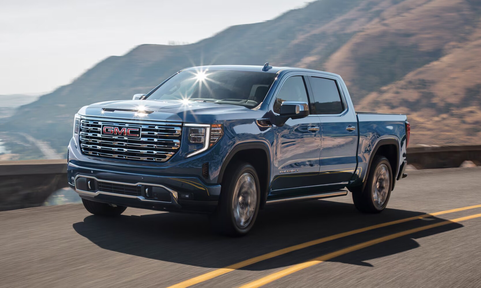 2026%20GMC%20Sierra%201500%20Denali%20-%20Octane%20GMC%20of%20Santa%20Fe.png Exterior front side angle image of a blue 2026 GMC Sierra 1500 Denali driving on a mountainous lakeside freeway