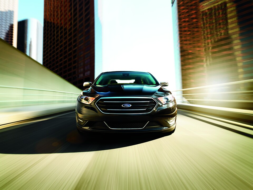 2017-Ford-Taurus-Limited-Sedan-L01.png black Ford Taurus driving towards the camera with tall city buildings on the side