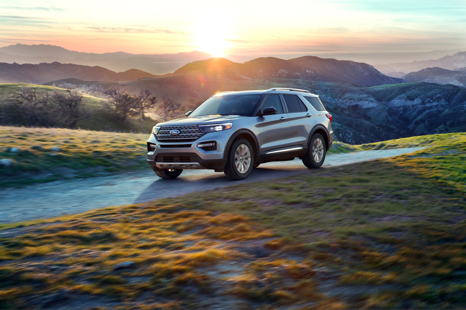 2021-Ford-Explorer-LTDhybrid-SUV-L02.png silver Ford Explorer hybrid SUV driving on a road next to rolling green hills