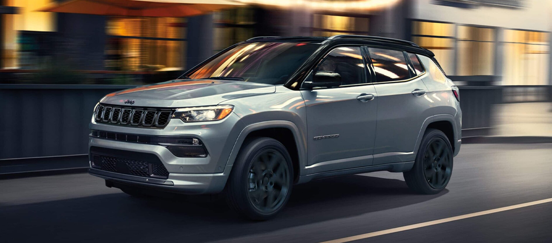 A silver Jeep Compass driving over a bridge in a city. A silver Jeep Compass driving over a bridge in a city.