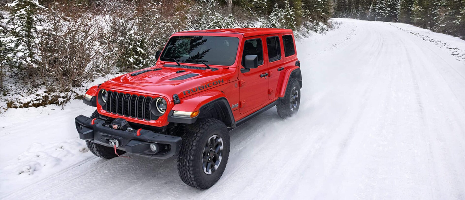A red 2026 Jeep Wrangler driving over a snow-covered road by a forest.