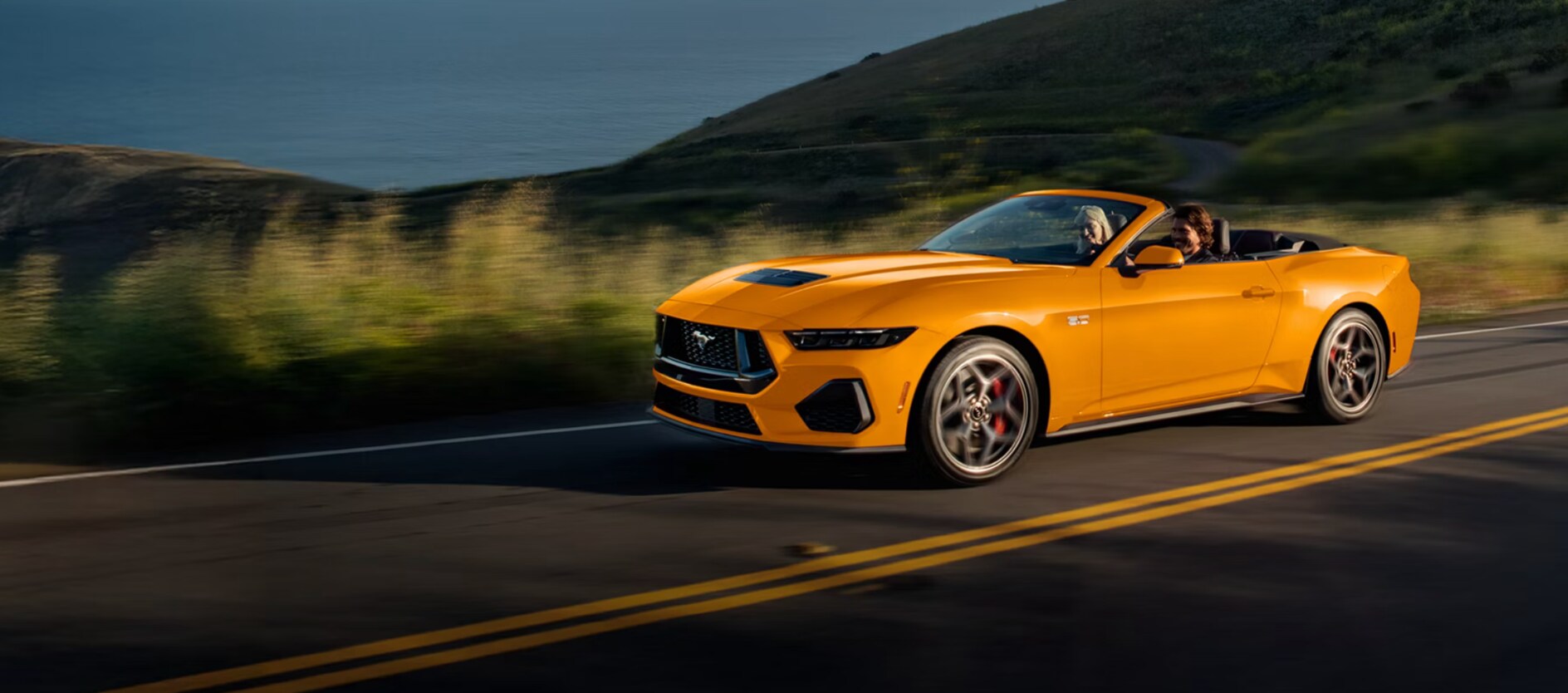 A yellow-orange Ford Mustang driving down the road by a hill.