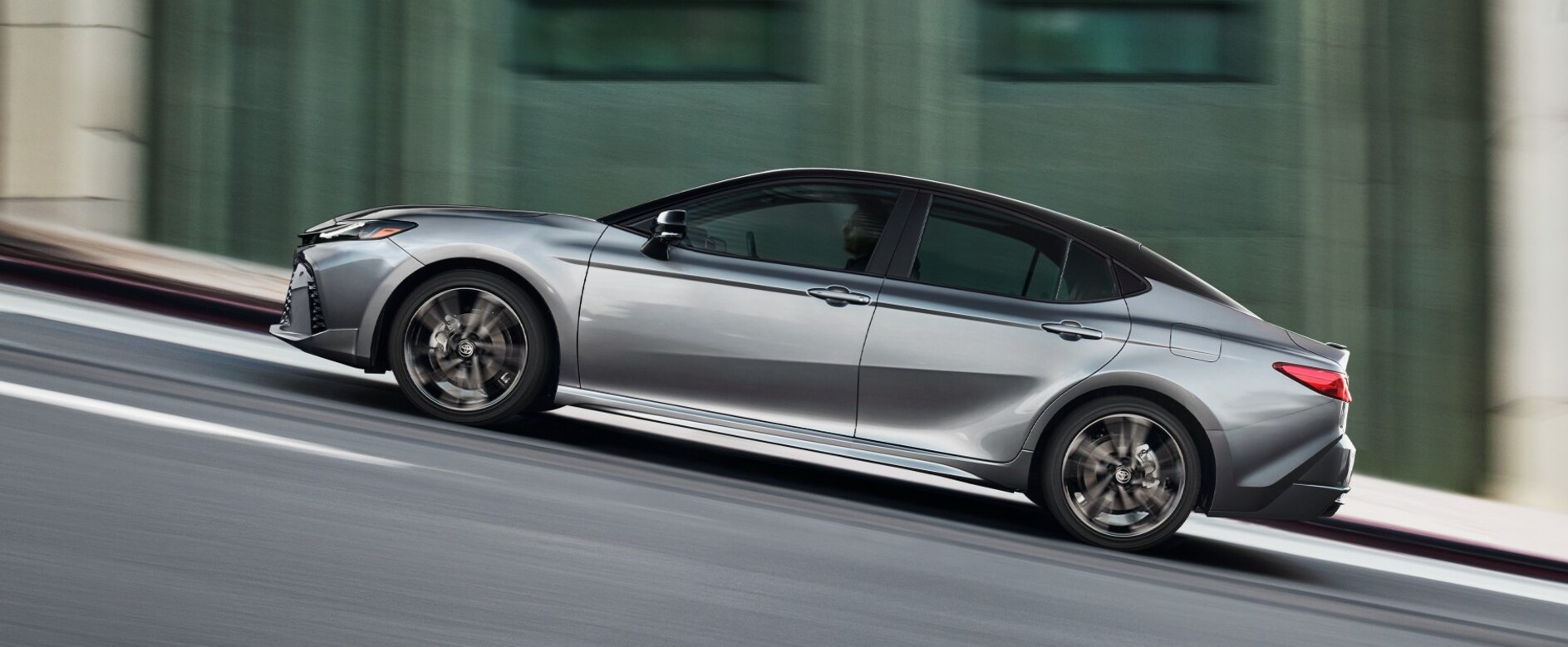 A silver 2026 Toyota Camry in profile driving up an inclined road. A silver 2026 Toyota Camry in profile driving up an inclined road.