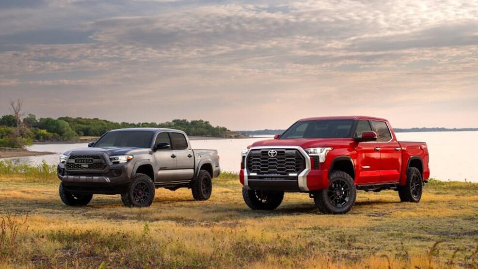 A gray Toyota Tacoma on a field by a lake, sitting next to a red Toyota Tundra.