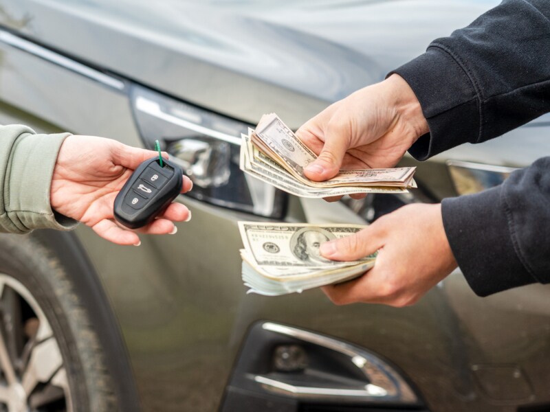 Two people exchange car keys and cash in front of a vehicle, suggesting a car sale. Hands are visible, conveying a transactional moment.