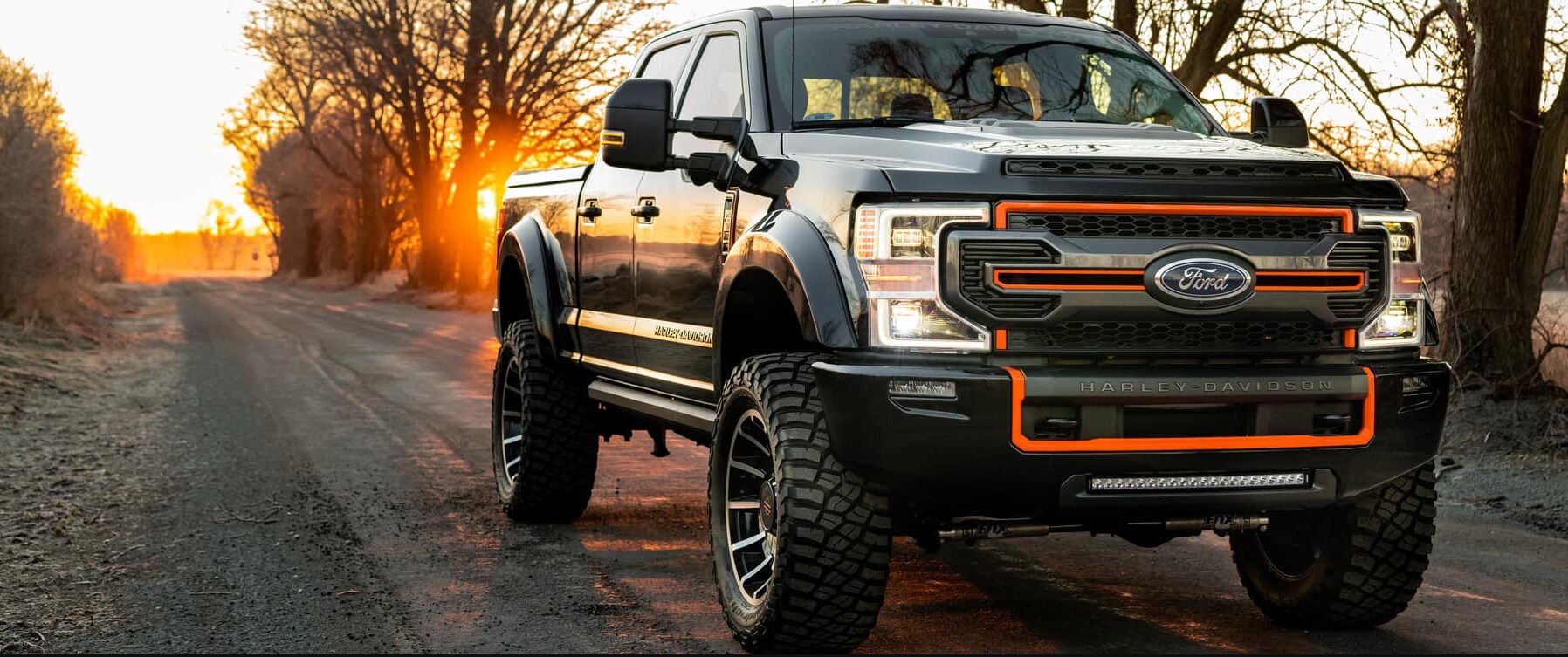 Ford F250 Super Duty Harley Davidson.PNG The sun is setting in the background of some leaf-less trees as a Black 2022 Ford F250 Super Duty Harley Davidson sits on a road facing the viewer, highlighting the black chassis, orange outlining on the front grille, and the words Harley Davidson embossed on the front