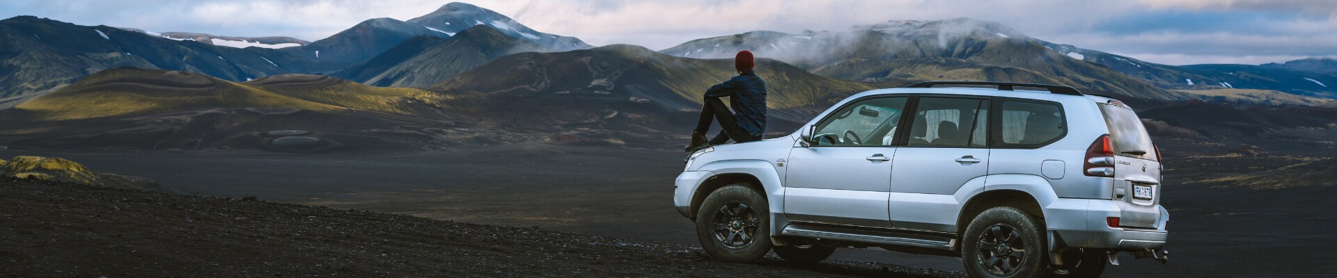 Best used AWD cars in Colorado at Phil Long Dealerships.png Driver side view of a used AWD car parked on a mountain side as the driver sits on the hood overlooking a beautiful mountain range in the distance
