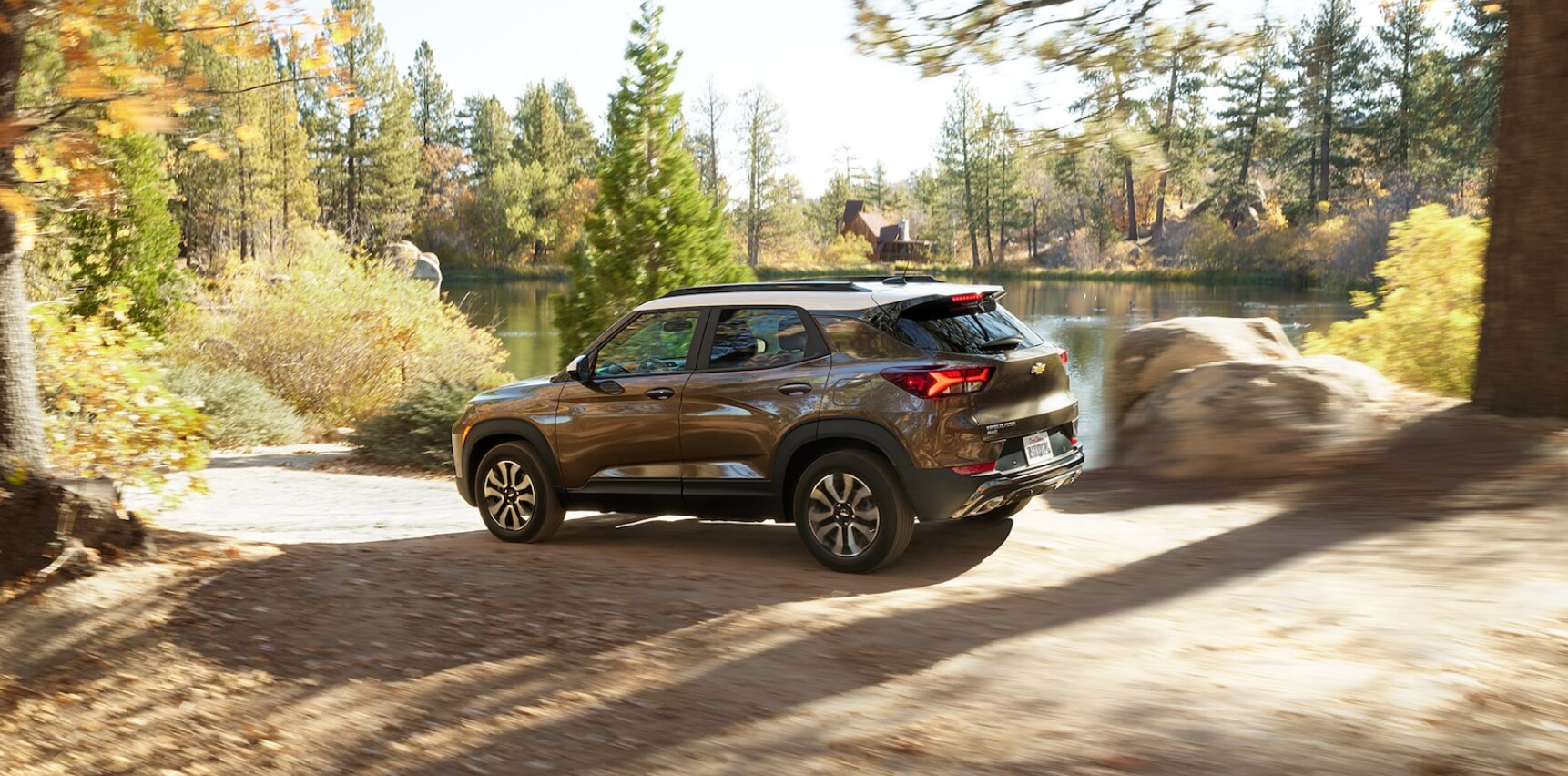 2022-Chevy-Trailblazer-2.PNG A crisp fall day and the sun shining through the leaves bordering a country road as a bronze metallic Chevy Trailblazer cruises past