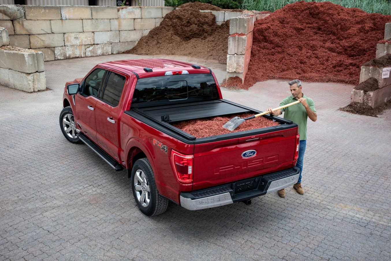 A cherry red F150 is parked at a construction site with the tonneau cover rolled back to reveal a load of mulch a man is shoveling out on the side