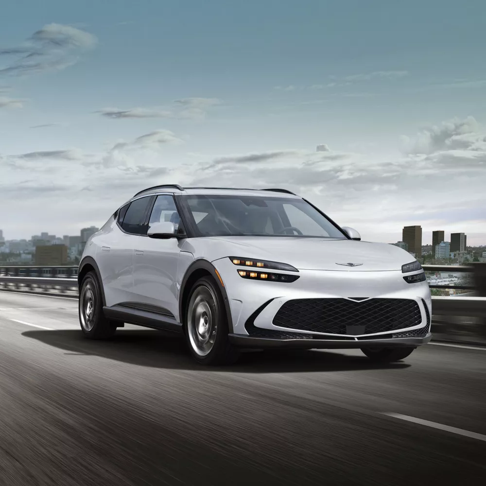 2025%20Genesis%20gv60.webp A new pearl white new 2025 Genesis GV60 driving on a highway during the day with a city in the background.