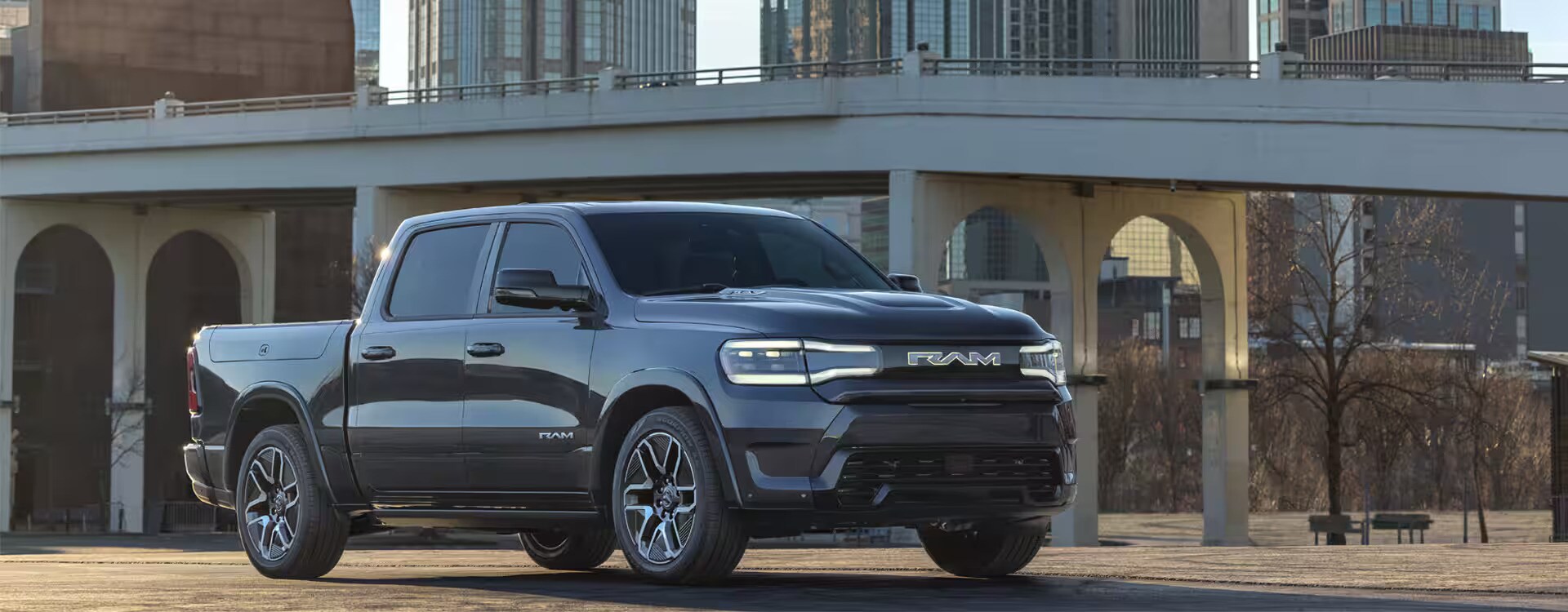 2025%20Ram%201500%20REV.jpg A new black 2025 Ram 1500 REV sitting idle with a bridge and a few city buildings in the background.