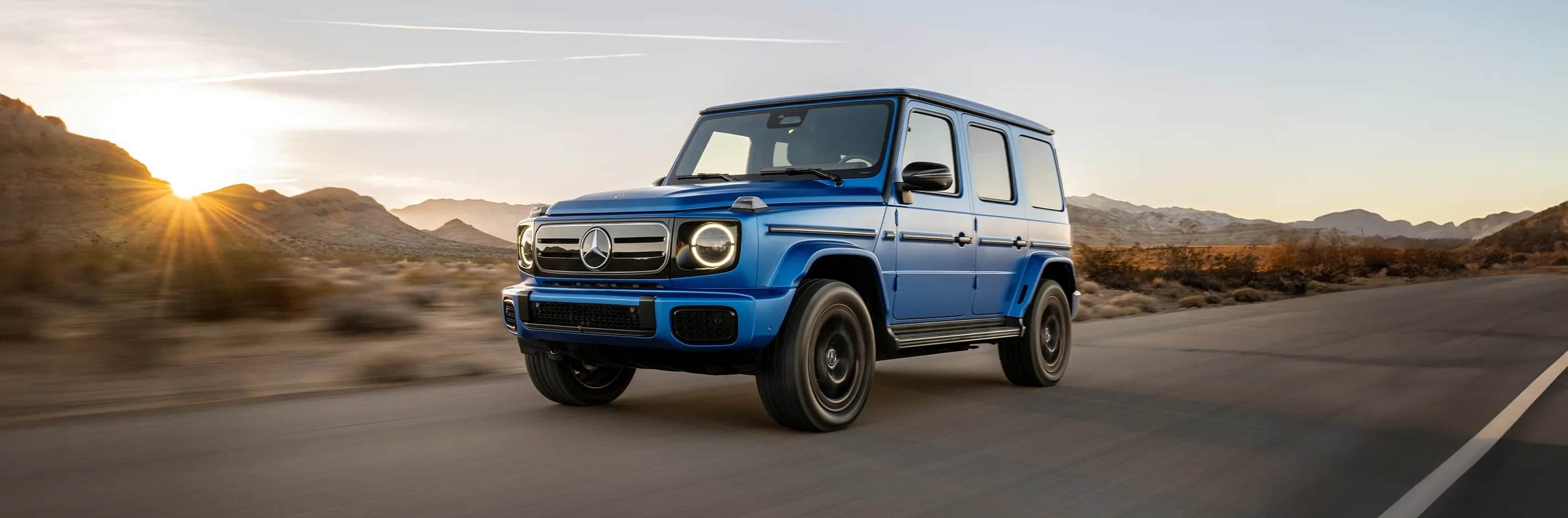 2025%20Mercedes%20Benz%20G%20580.webp A blue new 2025 Mercedes-Benz G-Class driving on a highway during the day in the dessert.