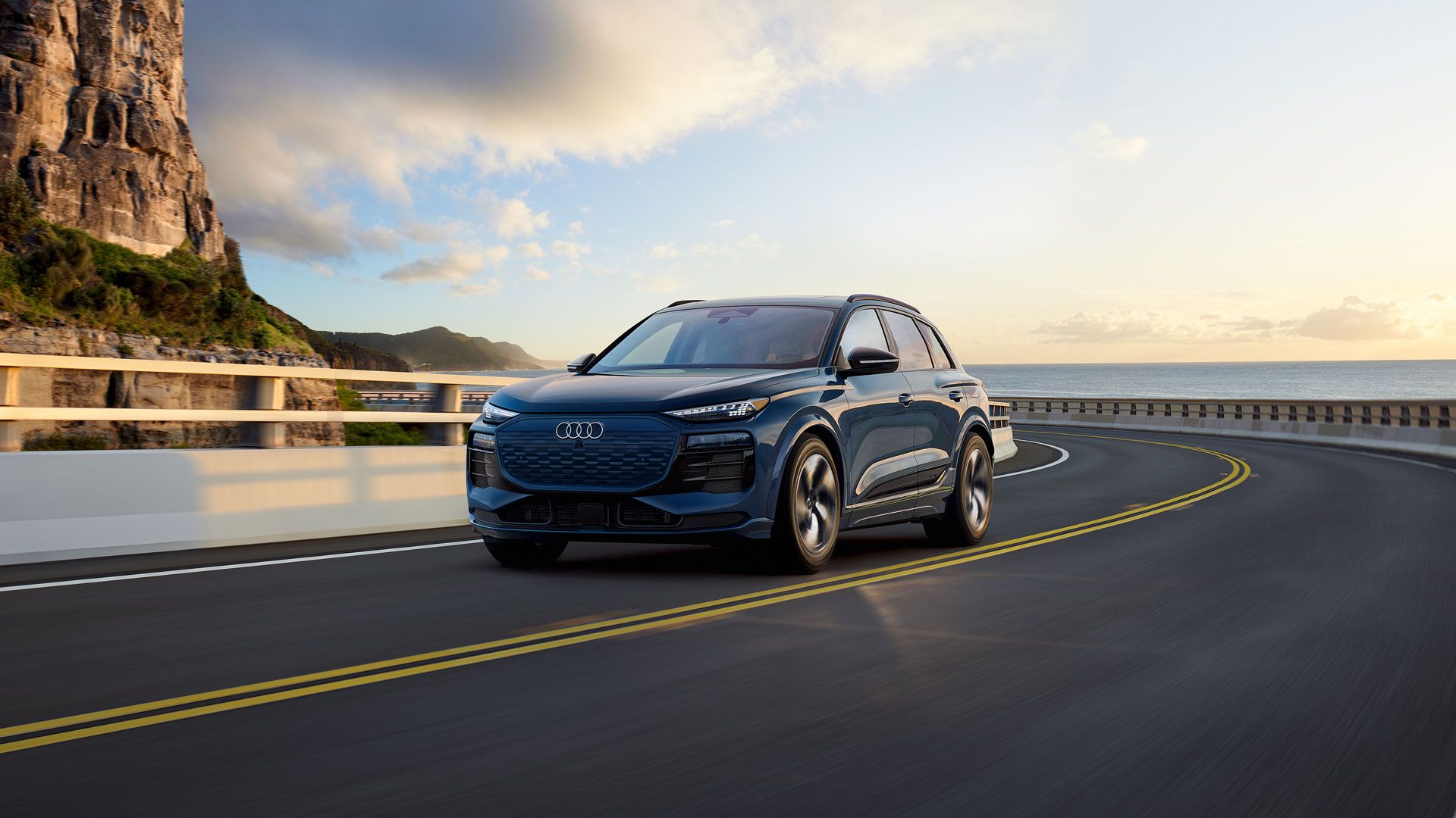 2025%20Audi%20Q6.jpg A new 2025 Audi Q6 E-Tron driving on a oceanside highway during the day.