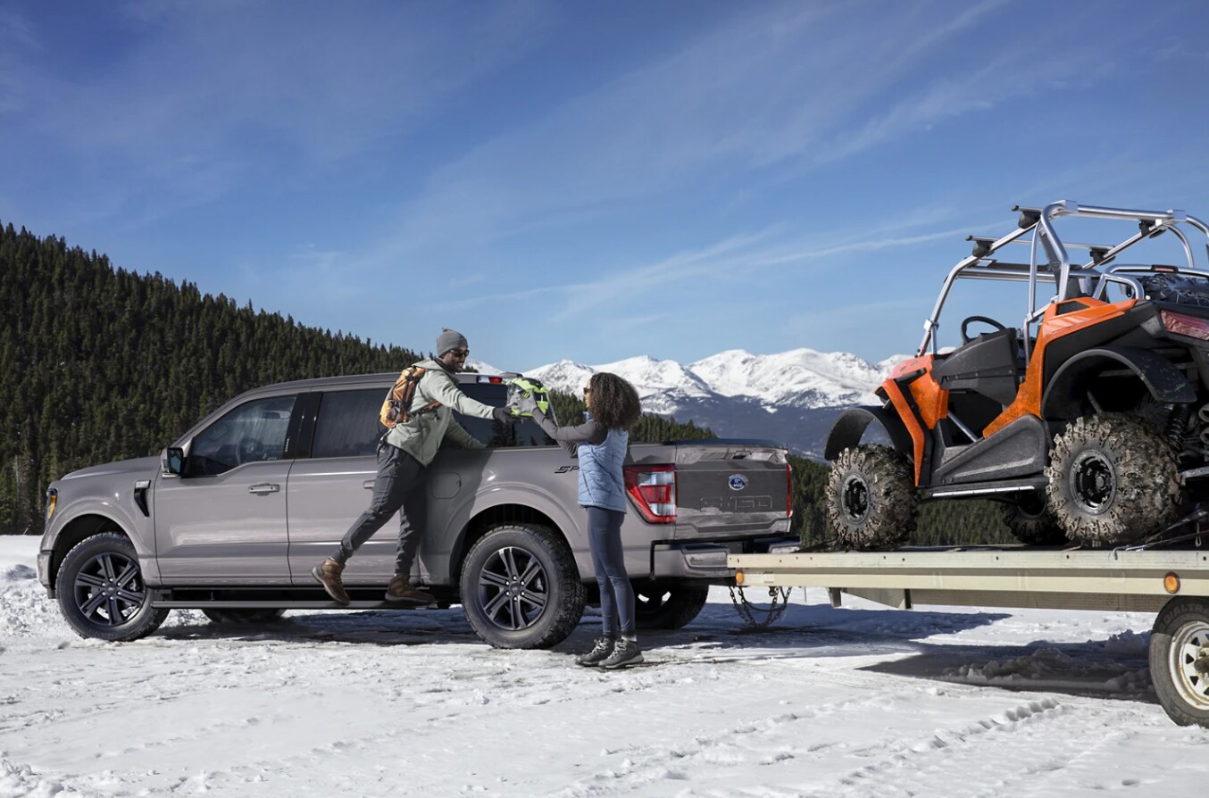 A silver F150 is towing a 4-wheeler and parked in a snow-covered scene with a backdrop of a dense evergreen forest and snow-capped mountain.  A man is standing on the side step of the car, handing something from the pickup bed to his wife
