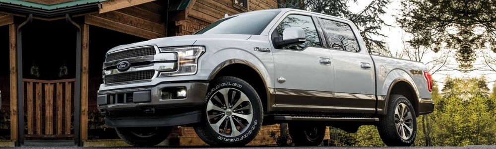 2019 Ford F-150 Truck in Denver at Phil Long Ford.png Ground level shot of the driver side of a white 2019 Ford F-150 truck parked outside a wooden cabin in the woods with a green roof