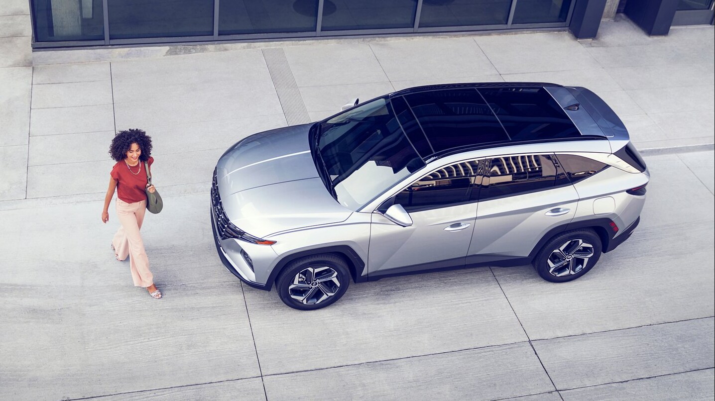 2022-Hyundai-Tucson-02.PNG A bird's eye view of a woman walking past the front of a silver Hyundai Tucson with a black glass panoramic sunroof parked in a city on a concrete pad