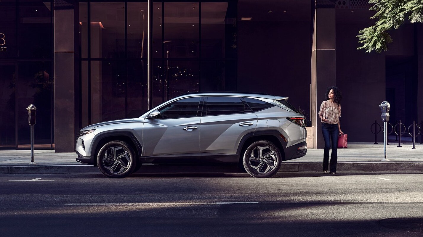 2022 Hyundai Tucson-2.PNG A silver 2022 Hyundai Tucson sits in profile view, parked on a city street as a woman stands by the hatch ready to cross the street towards the viewer.