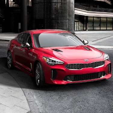2019kiastingerincoloradospringsatphillongkia The front angle of a shiny red 2019 Kia Stinger as it makes a right turn on an empty city street in front of a big industrial complex