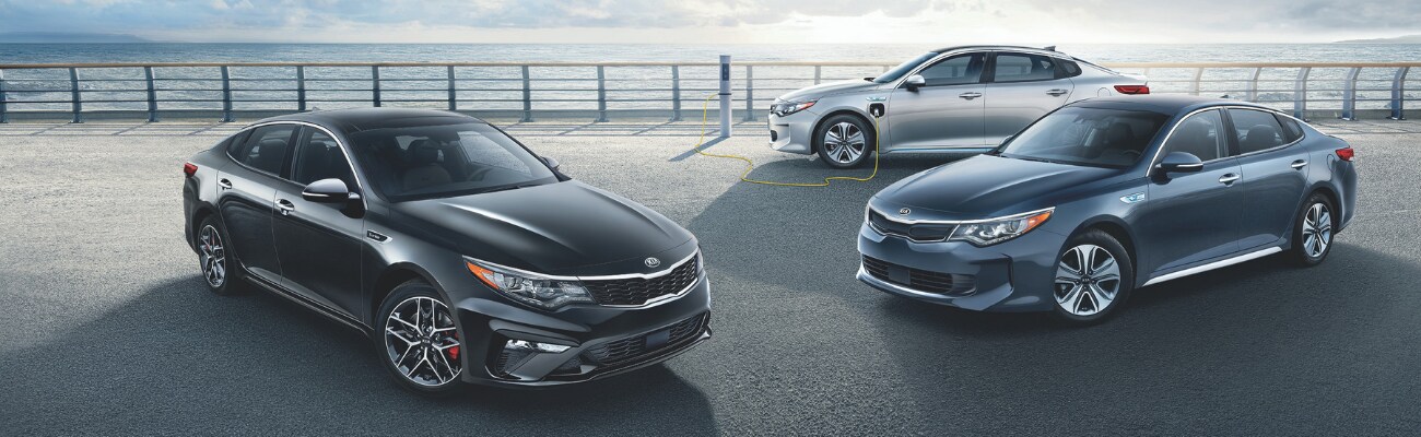 Zoomed out view of a 2019 Kia Optima, 2019 Kia Optima Hybrid, 2019 Kia Optima Hybrid Plug-In parked on a concrete ledge overlooking a body of water under the rising sun
