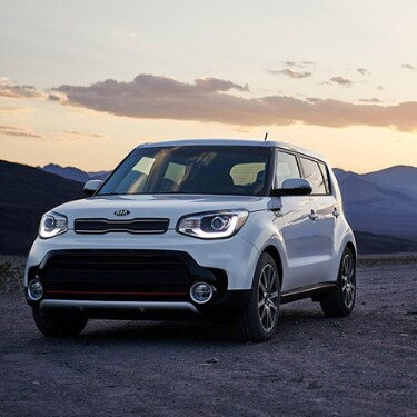 2019kiasoulincoloradospringsatphillongkia The front driver side of a white 2019 Kia Soul parked on a dirt path as the automatic headlights turn on with the sun setting behind a mountain range in the background
