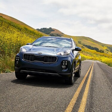 2019kiasportageincoloradospringsatphillongkia A view of the front of a steel blue 2019 Kia Sportage SUV as it casually drives down an empty country road through foothills cover with yellow dandelions