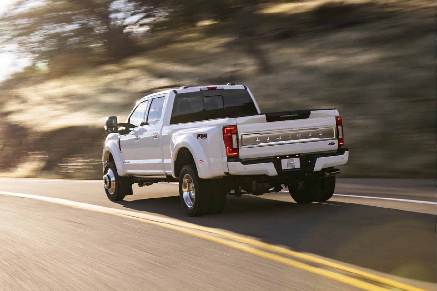 Ford-Super-Duty-2.PNG A white 2022 Ford Super Duty drives away from the viewer showing the profile and tailgate of the vehicle on a sunny day on a curving road past a blurred background
