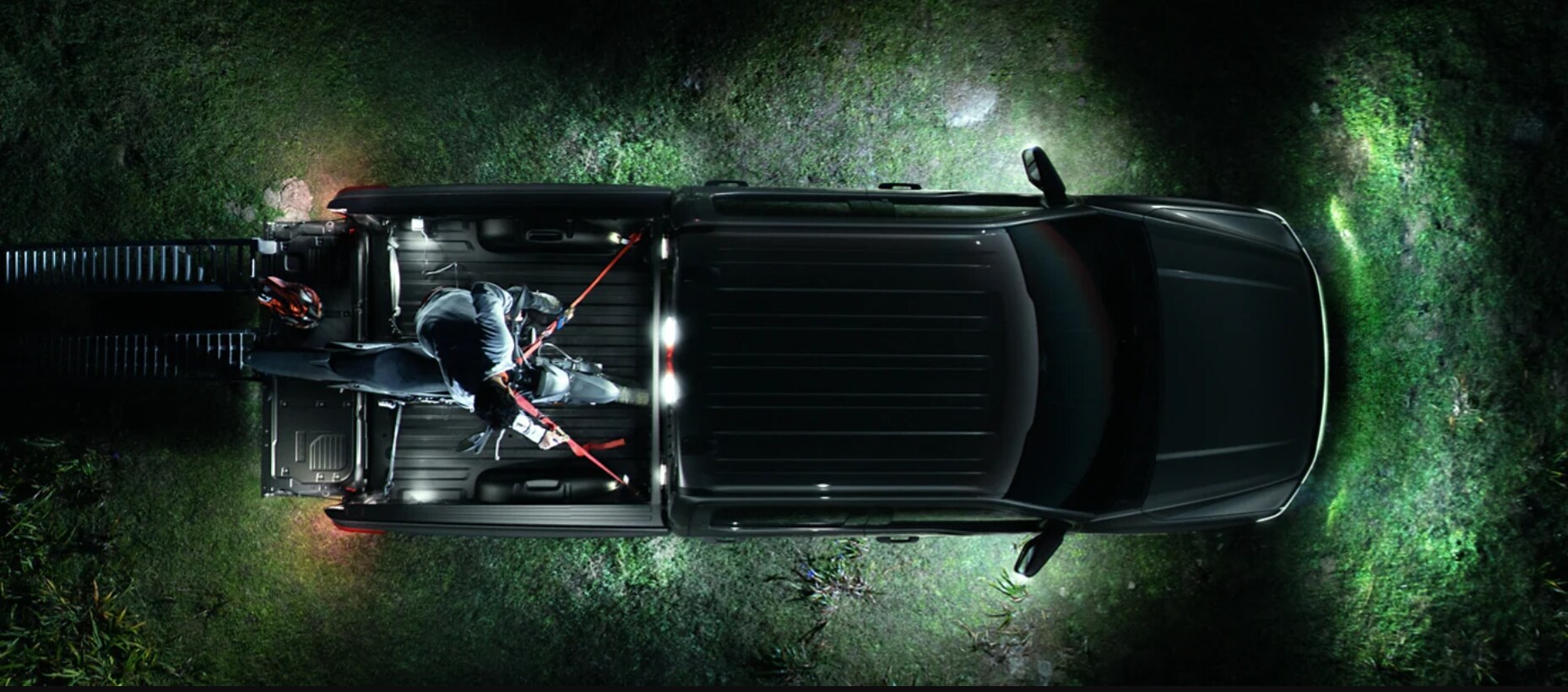 2022-Ford-F-150-Lightning-15.PNG An overhead view of the Ford F-150 parked at night in a field as a dirt bike driver loads the bike in the bed of the truck using metal ramps