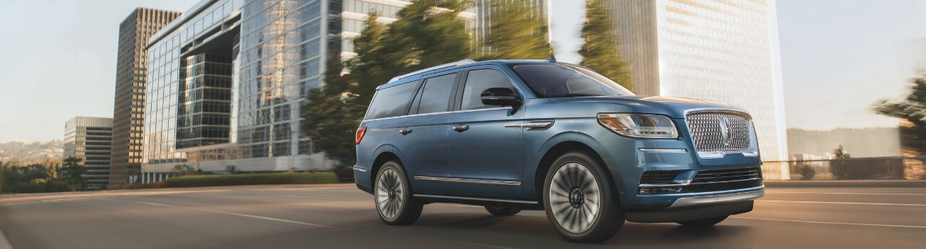 2019 Lincoln Navigator in Colorado Springs at Phil Long Lincoln.png View of the passenger side of a light blue 2019 Lincoln Navigator driving down a highway with blurred images of trees and tall city buildings in the background