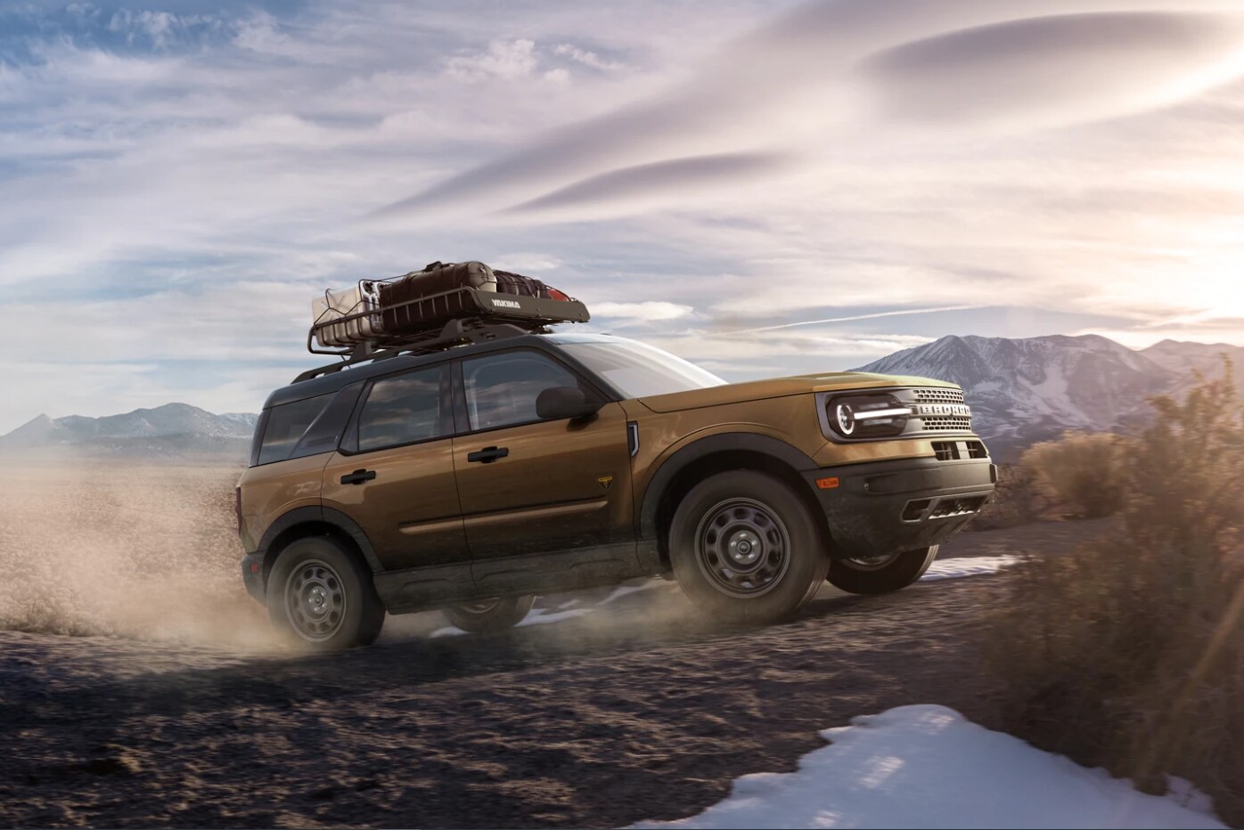 2022 Ford Bronco Sport Main 3.png A bronze metallic 2022 Ford Bronco Sport climbs a dirt hill with a rooftop rack full of equipment and kicks up a cloud of dust while the sun sets in the background in front of a mountain range