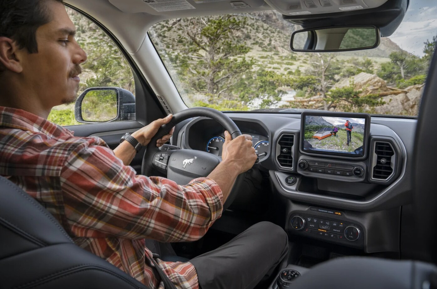 2022 Ford Bronco Sport Dash 1.png A mustachioed man in a plaid shirt sits in the driver's seat of a 2022 Ford Bronco Sport and stares at the view of the rearview camera on the center dash-mounted infotainment system.