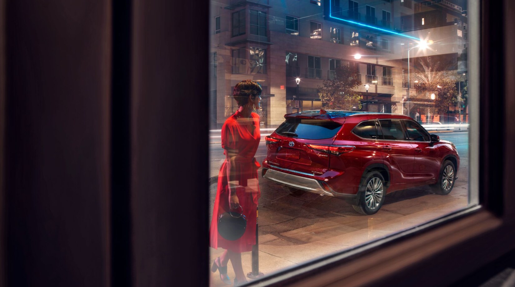 2022-Toyota-Highlander-Hybrid-2.PNG A view through a city window looking at a parked red 2022 Toyota Highlander and a woman standing behind it in a red dress admiring it