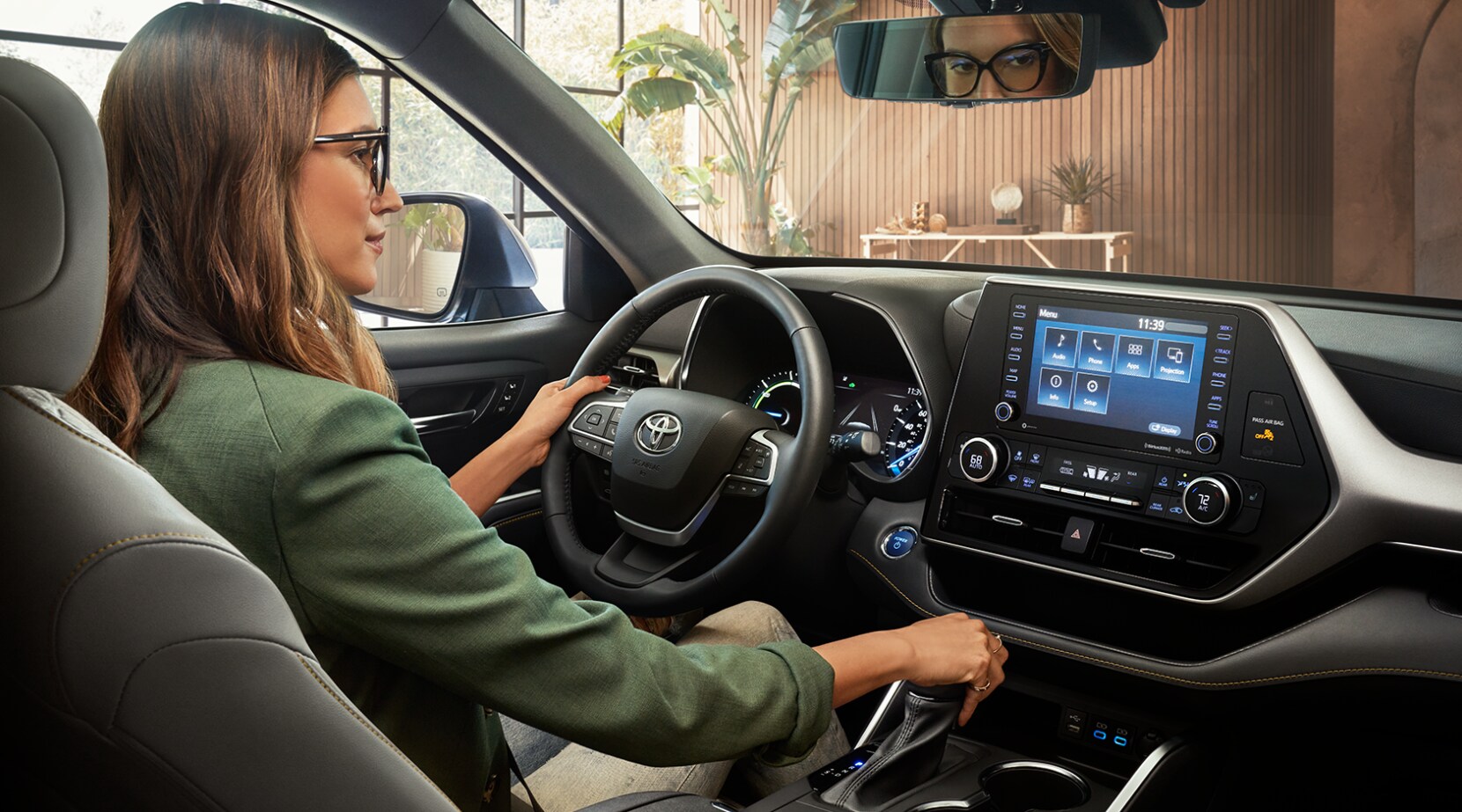 2022-Toyota-Highlander-Hybrid-4.PNG A woman sits in the driver's seat of a 2022 Toyota Highlander and looks at the large infotainment screen mounted in the middle of the black dash with silver accents