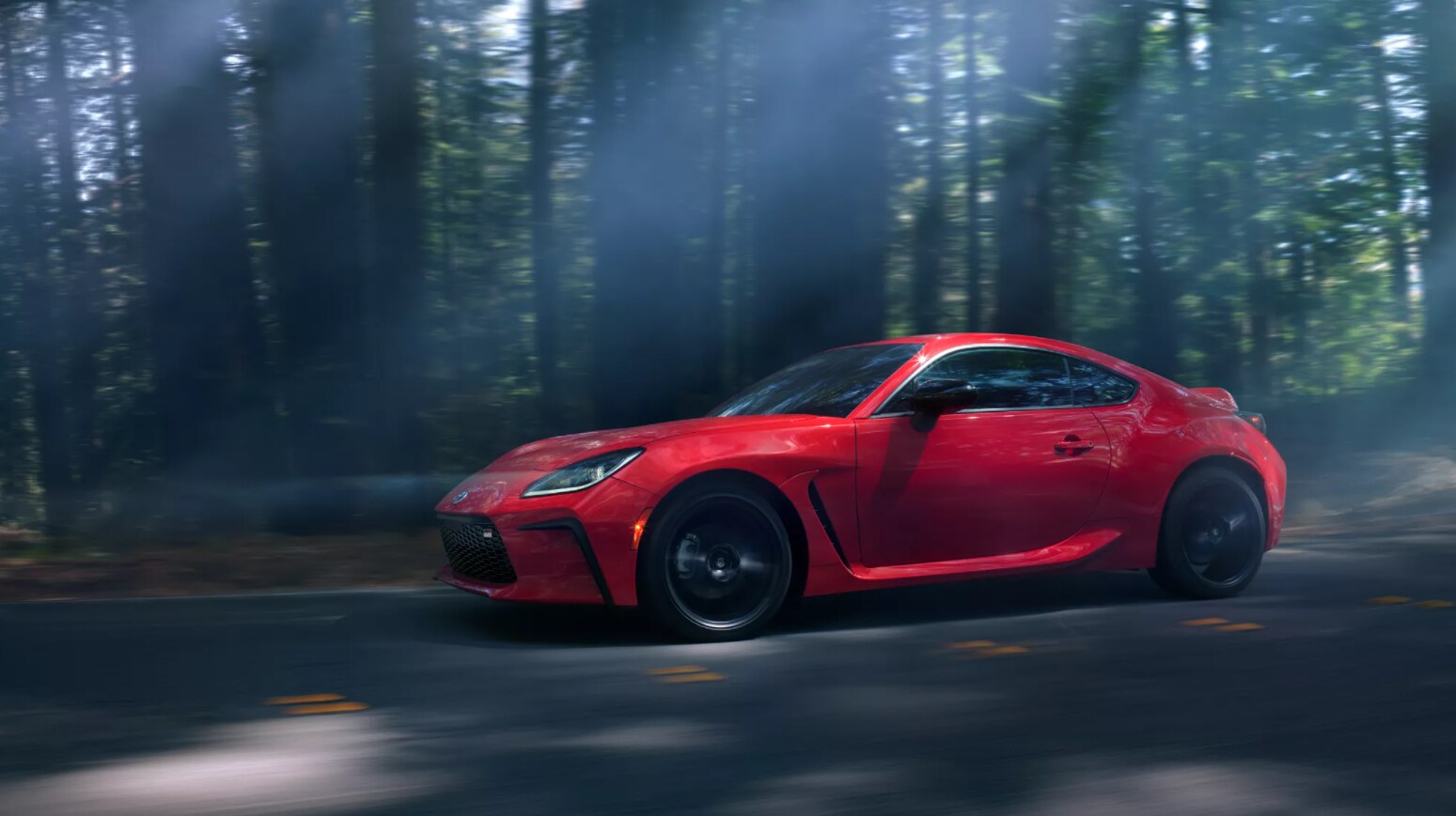 2022-Toyota-GR86-03.PNG A shiny classic red Toyota GR86 speeds along a winding road in a shadowy forest