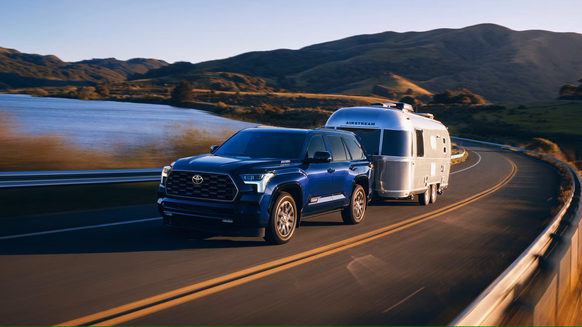 Metallic dark blue 2025 Toyota Sequoia hauling an airstream on a road over a lake.