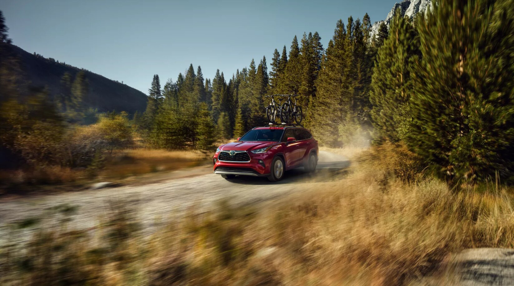 2022-Toyota-Highlander-Hybrid-1.PNG A red 2022 Toyota Highlander Hybrid with mountain bikes mounted to the roof speeds down a gravel road leaving a trail of dust behind as it cruises through a mountain forest during the day.