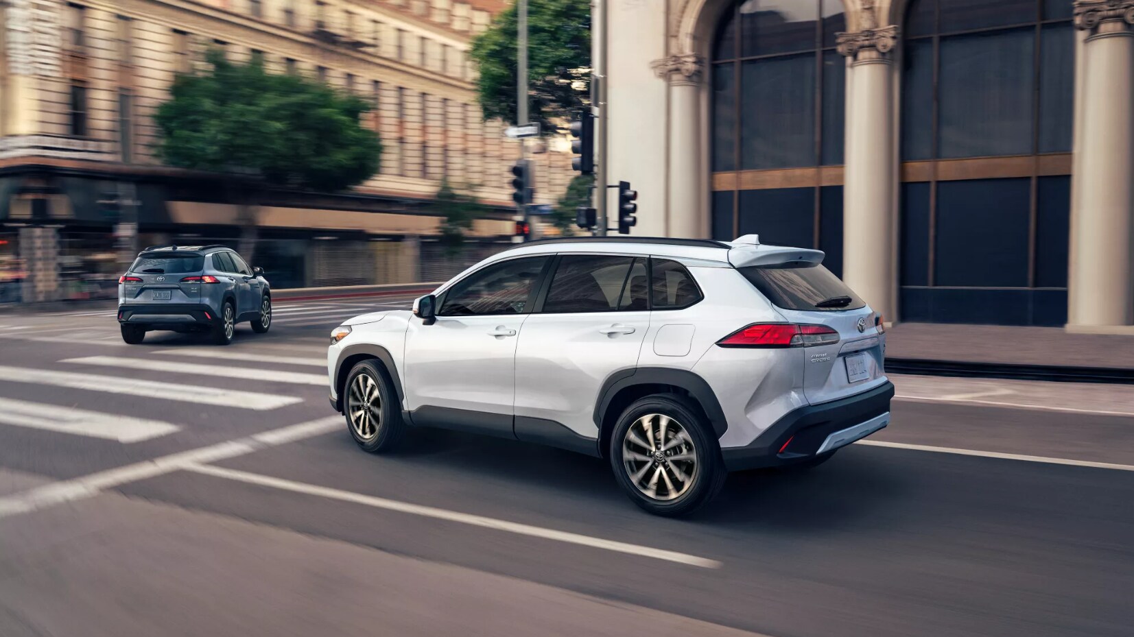 2022-Toyota-Corolla-Cross-3.PNG A silver 2022 Toyota Corolla Cross in profile view passes the viewer as it drives down a city street about to cross an intersection during the day.