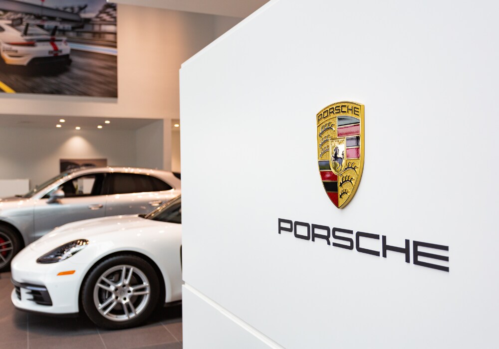porsche-dealership-charlottesville-va.png The Porsche logo inside the Porsche Charlottesville dealership. There are Porsche cars parked in the background