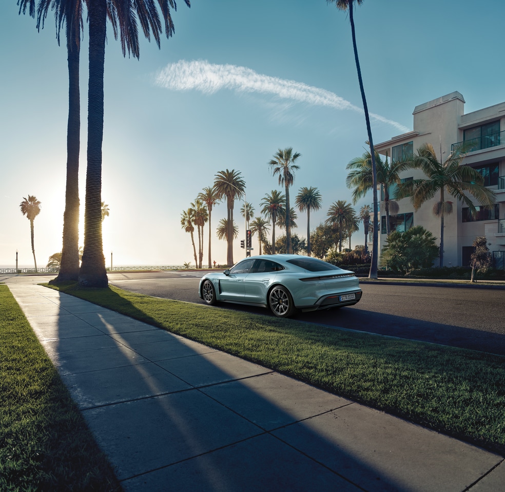 light blue Porsche Taycan parked on a palm tree lined street, next to an apartment building