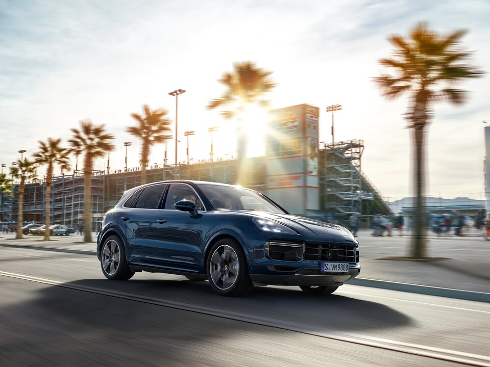 2019-Porsche-Cayenne-Turbo-SUV-L01.png dark blue Porsche Cayenne SUV driving by a palm tree lined street, next to a stadium