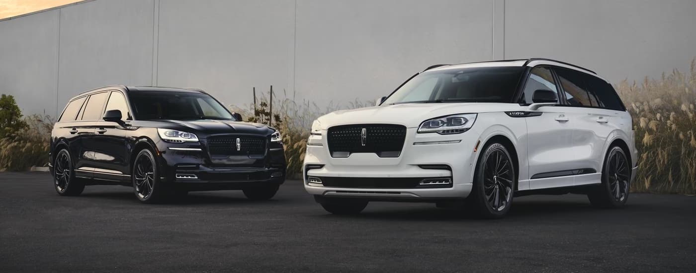 Lincoln Aviator for Sale - 2024 Lincoln Aviator - Black and White (1).jpg A black and a white 2024 Lincoln Aviator are shown parked near a tall concrete wall.