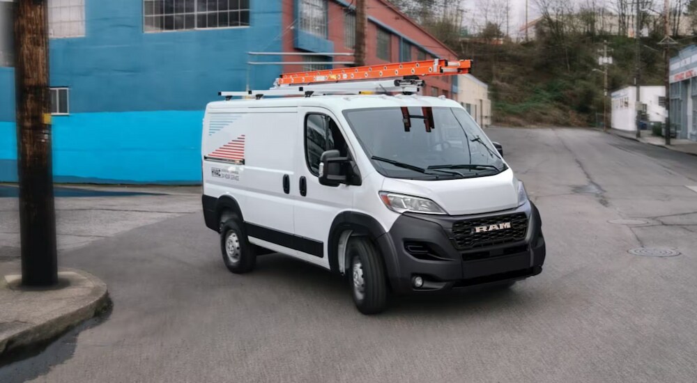 A white 2024 Ram ProMaster Cargo Van with ladders on the roof driving around a corner.