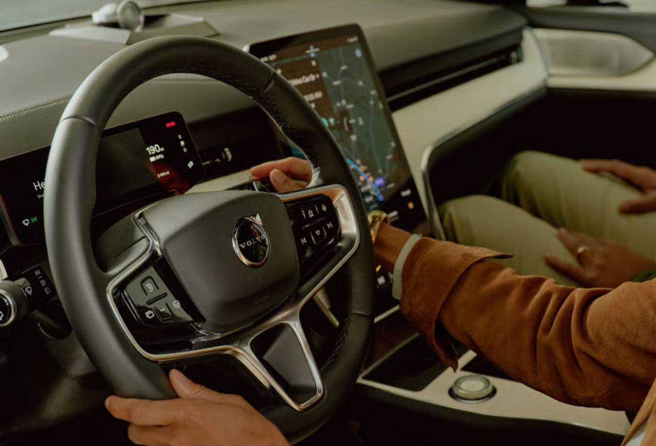 Close-up of a Volvo EX90 interior showing a person holding the steering wheel with a touchscreen displaying a map.