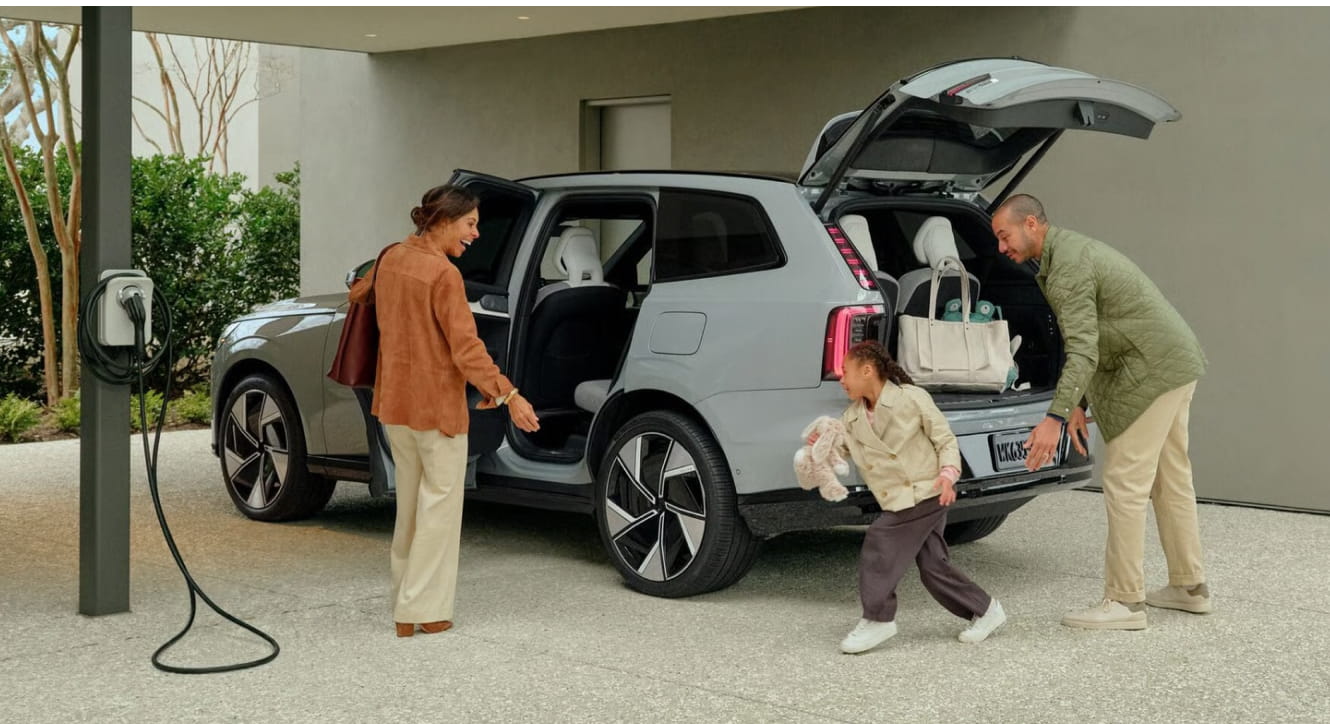 A family prepares for a trip with their Volvo EX90 SUV. The car is parked, charging, with the trunk open.
